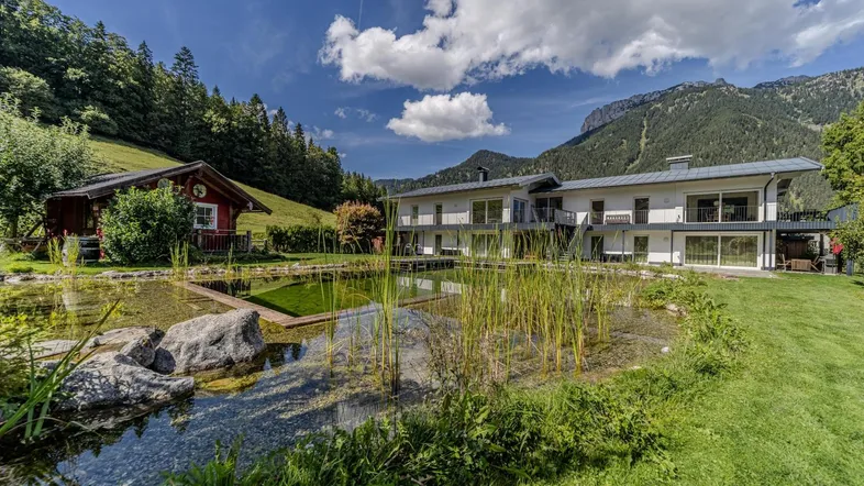 Großzügige Außenanlage mit Naturteich, umgeben von grünen Wiesen und Bergen.