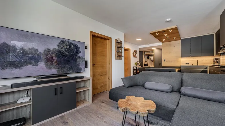 Modernes Wohnzimmer mit großem Fernseher, bequemer Couch und offener Küche im Hintergrund.