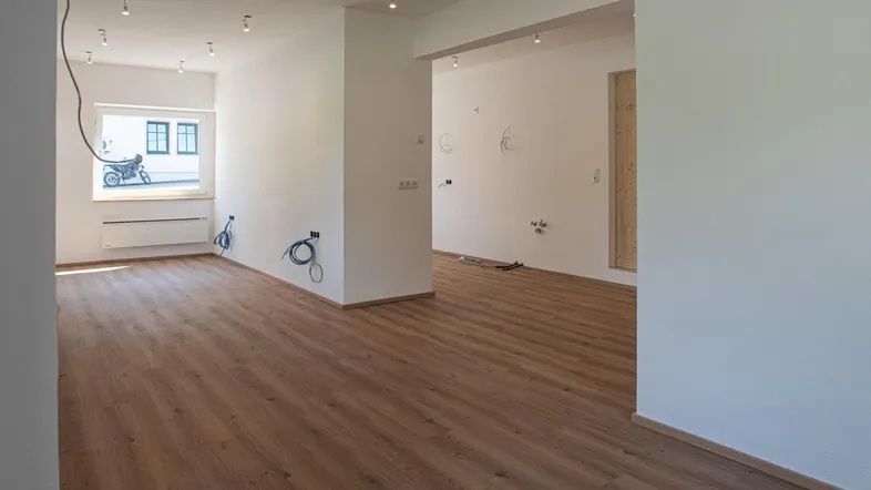 Leerer Innenraum in einem Neubau mit Laminatboden, weißen Wänden und sichtbaren Elektroinstallationen.