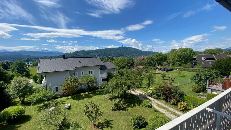 Weitläufiger Blick vom Balkon über die grüne Umgebung mit einem Haus und Bergen im Hintergrund.