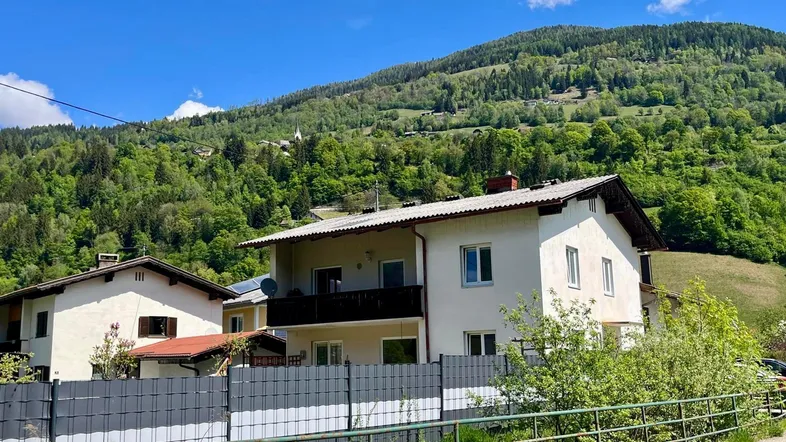 Weißes Wohnhaus mit Balkon und Satteldach vor einer bewaldeten Berglandschaft.