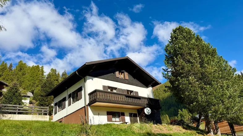 Charmantes Haus im alpinen Stil mit Holzbalkon und Satteldach, eingebettet in eine grüne Hügellandschaft.