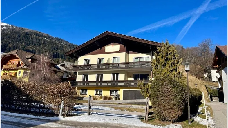 Mehrstöckiges Wohnhaus in ländlicher Umgebung mit Balkonen und Bergblick, umgeben von Bäumen und Schnee.
