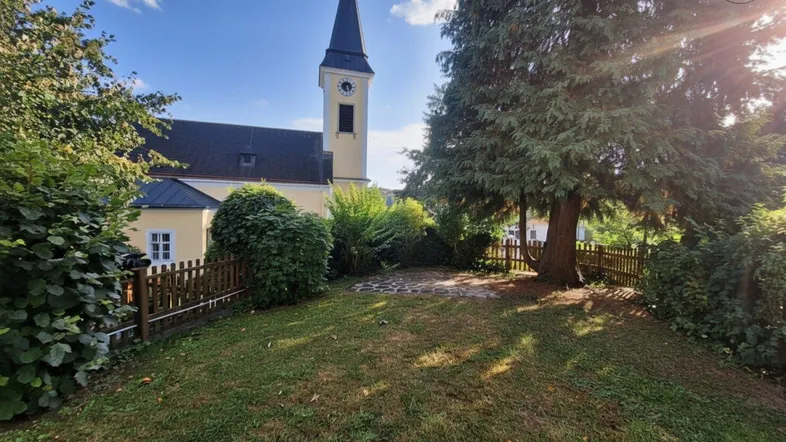 Gepflegter Gartenbereich mit Rasenfläche, Bäumen und Blick auf die nahegelegene Kirche unter blauem Himmel.
