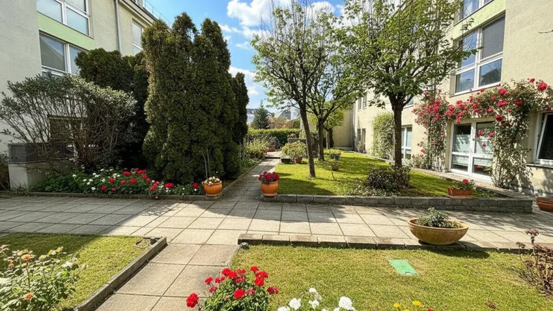 Sonnige Gartenanlage mit gepflasterten Wegen, bunten Blumenbeeten und hohen Bäumen.