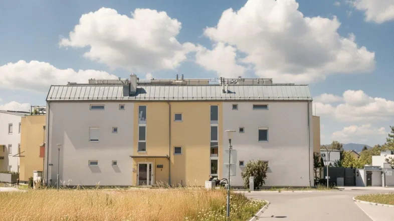 Mehrfamilienhaus mit moderner Fassade und umliegender Grünfläche unter blauem Himmel.