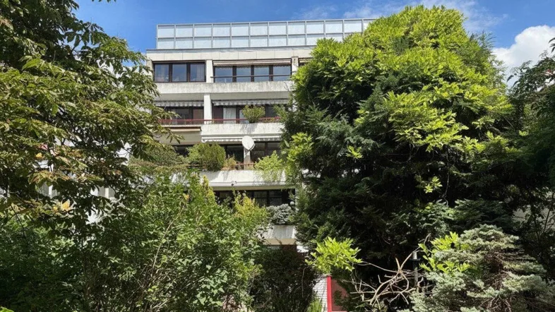 Mehrfamilienhausfassade umgeben von üppigem Grün und Bäumen unter blauem Himmel.