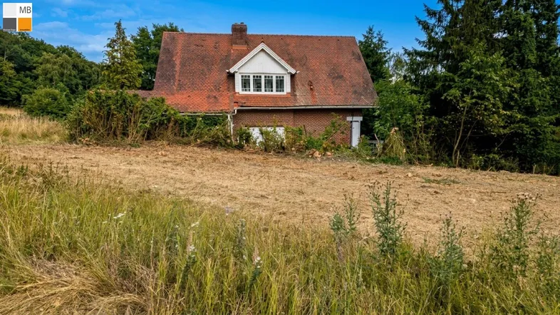 Altes Backsteinhaus mit rotem Ziegeldach und verwildertem Garten auf einem großen Grundstück.