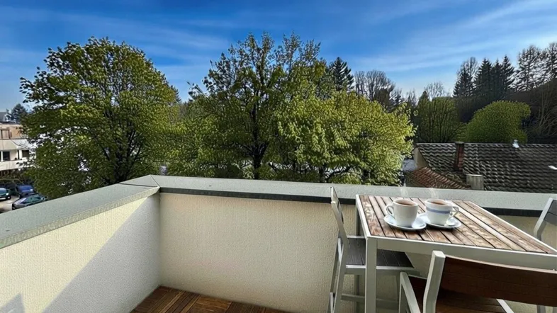 Gemütlicher Balkon mit Holzboden und Sitzgelegenheiten, umgeben von grünen Bäumen unter blauem Himmel.