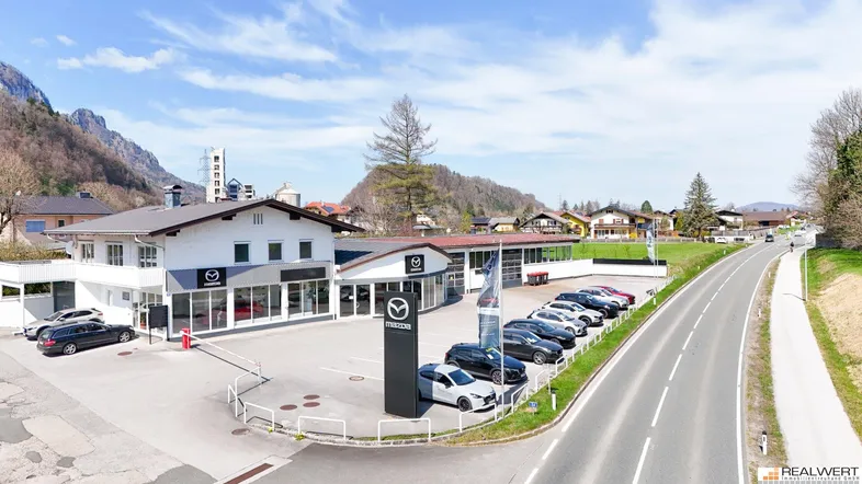 Luftaufnahme eines Autohauses mit Parkplätzen und umliegender Berglandschaft unter blauem Himmel.