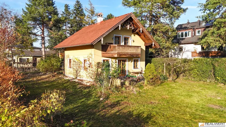 Traditionelles Einfamilienhaus mit gelber Fassade, Holzbalkon und großem Gartenbereich unter blauem Himmel.