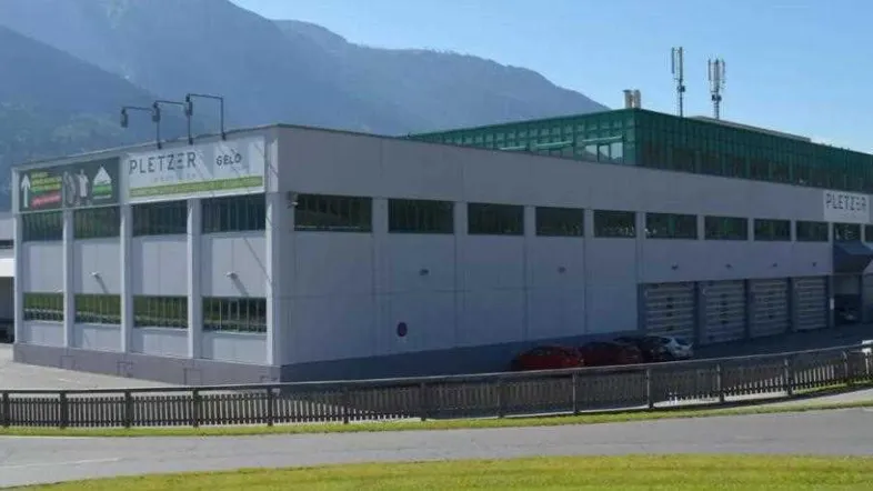 Großes Industriegebäude mit moderner Fassade, Laderampen und Parkplätzen vor einer Bergkulisse.