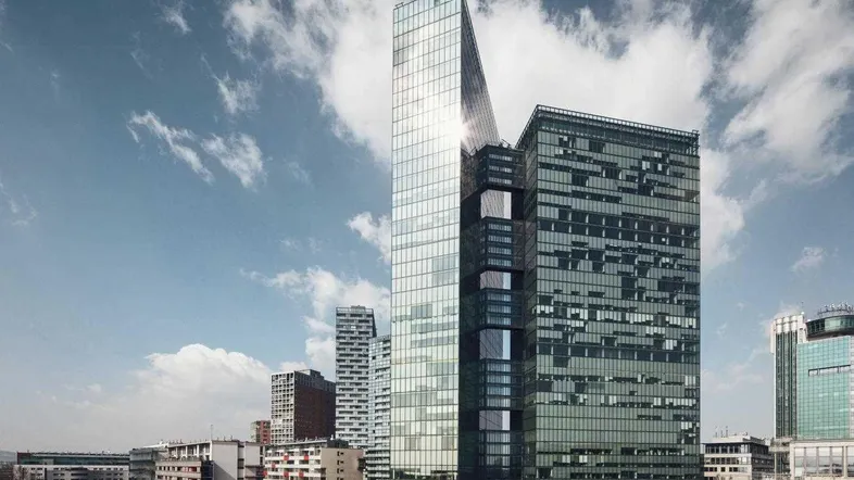 Moderne Glasfassade eines Hochhauses unter blauem Himmel mit Wolken, umgeben von städtischer Architektur.