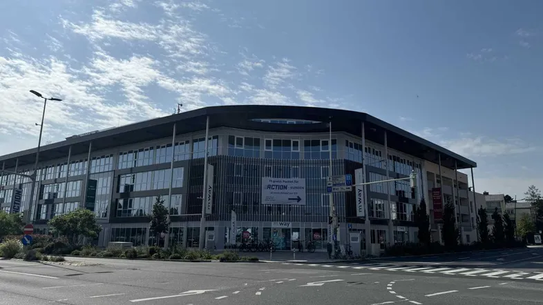 Modernes Geschäftsgebäude an einer Kreuzung mit großen Glasflächen und Werbebannern unter blauem Himmel.