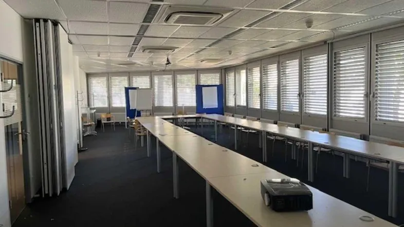 Großer Konferenzraum mit U-förmiger Tischordnung, vielen Fenstern mit Jalousien und einem Beamer.