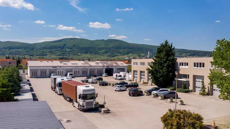 Luftaufnahme des Gewerbeobjekts mit großem Parkplatz und mehreren Gebäuden vor einer Bergkulisse.