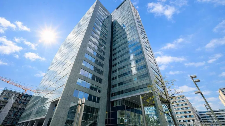 Modernes Hochhaus mit markanter Glasfassade und Betonstrukturen, aufgenommen an einem sonnigen Tag unter blauem Himmel.