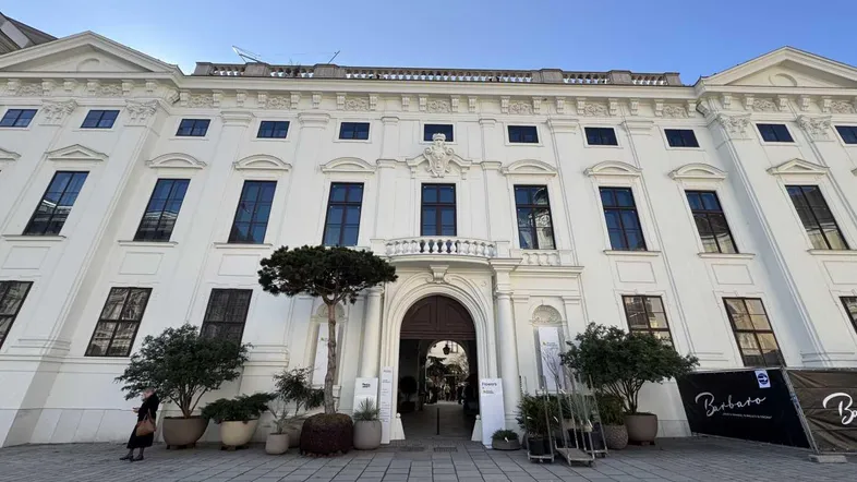 Klassisches weißes Gebäude mit vielen Fenstern, einem großen Torbogen und einem kleinen Balkon über dem Eingang.