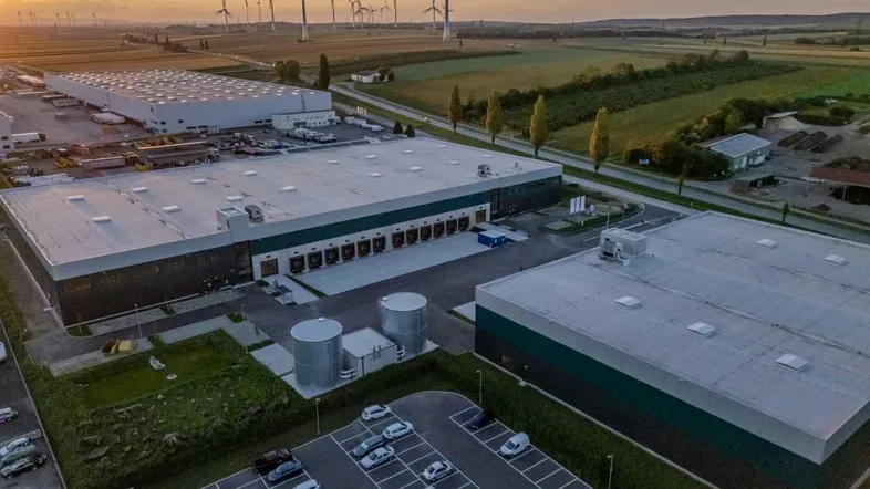Luftaufnahme eines modernen Logistikparks mit mehreren Lagerhallen, Parkplätzen und Windkraftanlagen im Hintergrund bei Sonnenuntergang.