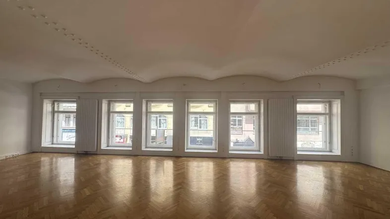 Geräumiger, heller Raum mit Fischgrätparkett und einer Reihe großer Fenster, die viel Tageslicht hereinlassen.