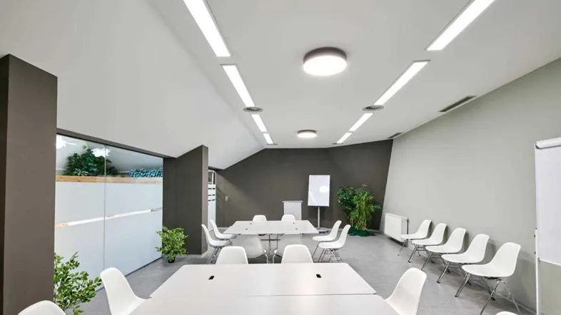 Moderner Konferenzraum mit weißen Tischen und Stühlen, linearen Deckenleuchten und einer Trennwand aus Milchglas.