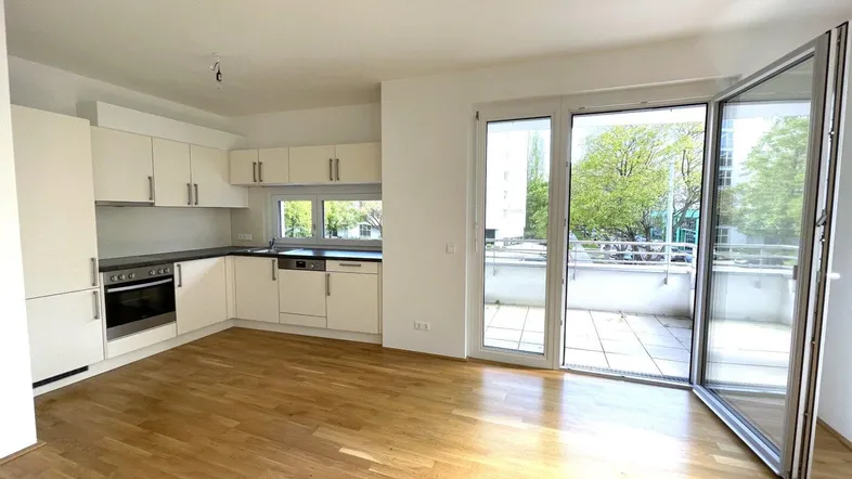 Heller offener Wohnbereich mit integrierter Küche, Holzboden und direktem Zugang zum Balkon.