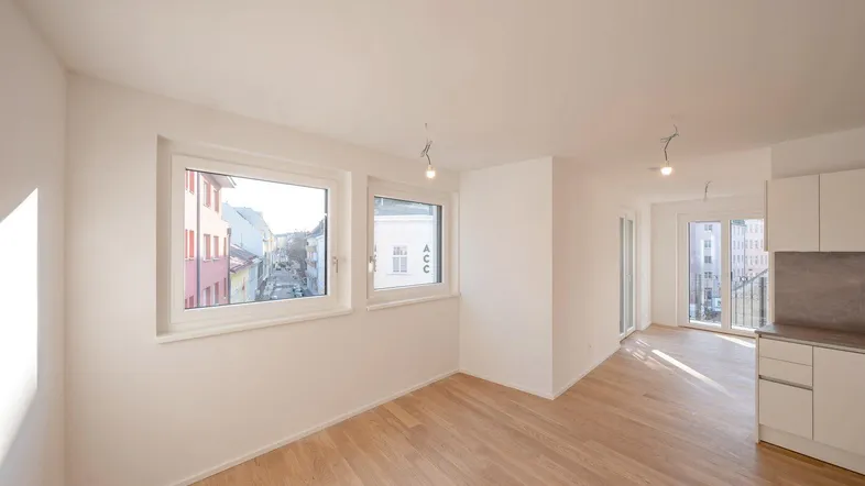 Helles Wohnzimmer mit Parkettboden, weißen Wänden und Blick auf die Straße, teilweise offene Küche sichtbar.
