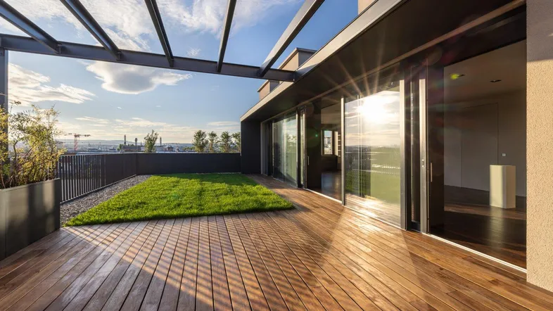 Große Dachterrasse mit Holzdeck, Grünfläche und Pergola, bietet einen weiten Blick über die Stadt bei Sonnenuntergang.