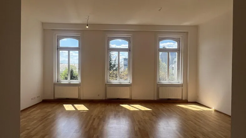 Helles, leeres Zimmer mit Parkettboden und drei hohen Bogenfenstern, die viel Tageslicht hereinlassen.