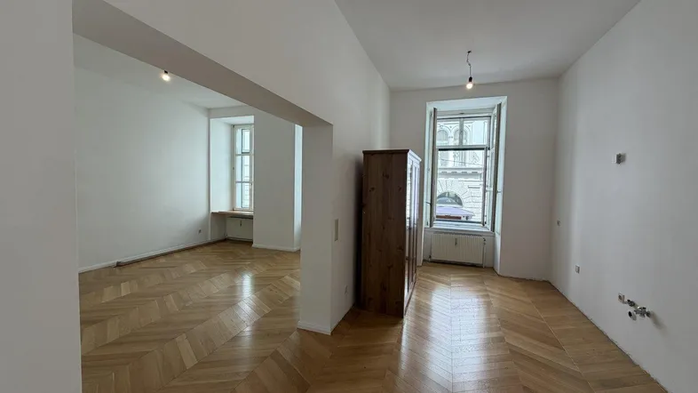 Helles Zimmer mit Fischgrätparkettboden und hohen Fenstern, das viel Tageslicht hereinlässt.