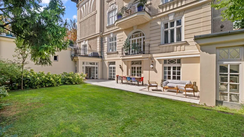 Großer Garten mit gepflasterter Terrasse und Sitzgelegenheiten vor einer eleganten Gebäudefassade.