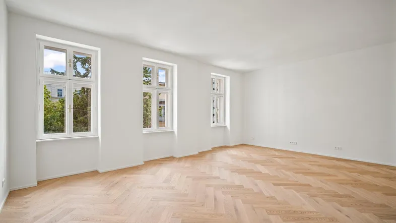 Helles, leeres Zimmer mit Fischgrätparkett und hohen Fenstern, die viel Tageslicht hereinlassen.