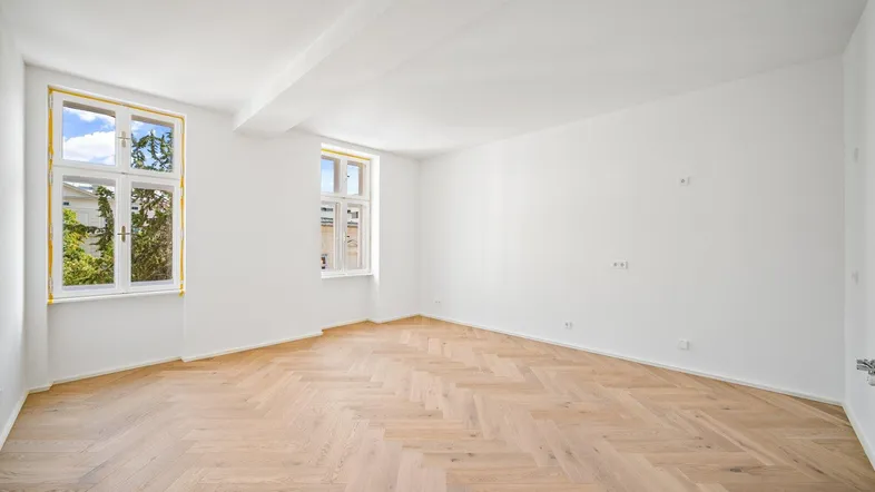 Helles Zimmer mit Fischgrätparkett und zwei hohen Fenstern, die viel Tageslicht hereinlassen.