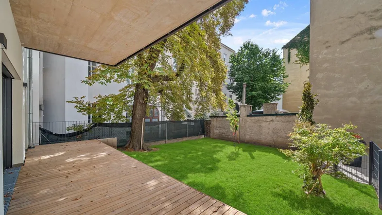Große Holzterrasse mit angrenzendem Rasenbereich und schattenspendenden Bäumen.