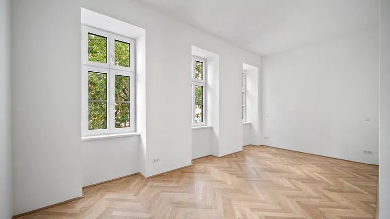 Helles Zimmer mit Fischgrätparkett und hohen Fenstern, das viel Tageslicht hereinlässt.