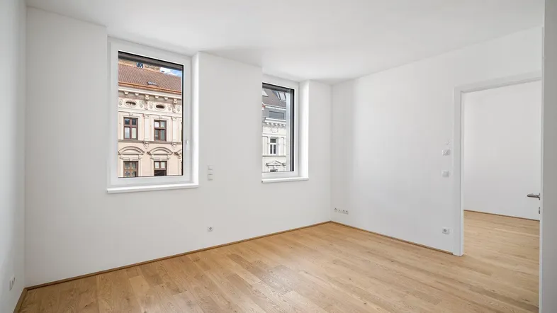 Helles, leeres Zimmer mit Holzboden und zwei Fenstern, die einen Blick auf die Stadt bieten.