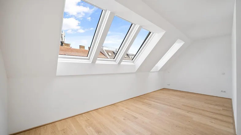 Helles Zimmer im Dachgeschoss mit Holzboden und mehreren Dachfenstern, die viel Tageslicht hereinlassen.