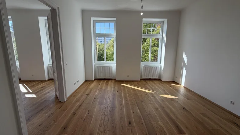 Helles Zimmer mit neuem Parkettboden, weißen Wänden und Blick ins Grüne durch zwei Fenster.