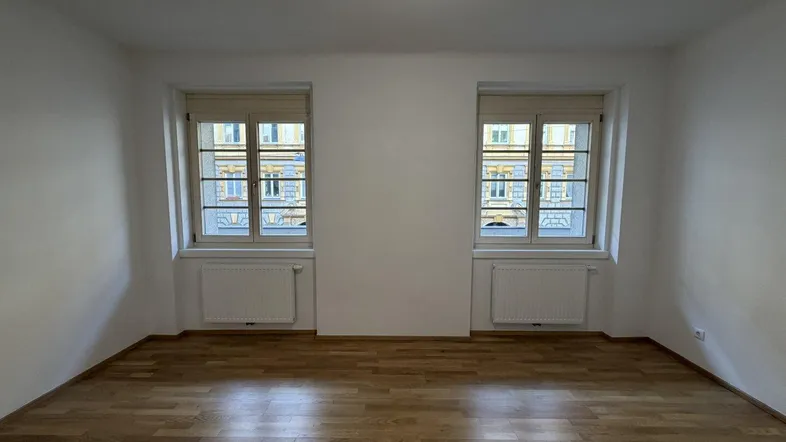 Helles Zimmer mit Parkettboden und zwei Fenstern, die viel Tageslicht hereinlassen.