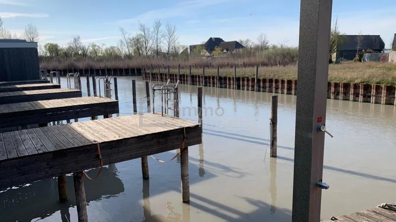 Anlegestelle mit Holzstegen und Außendusche am Wasser, umgeben von natürlicher Uferlandschaft.