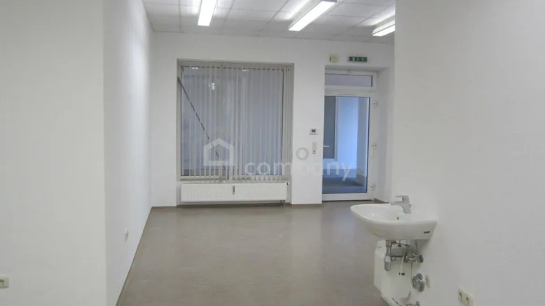 Leerer Raum mit weißen Wänden, grauem Boden, Fenster mit Jalousien und einem Waschbecken.