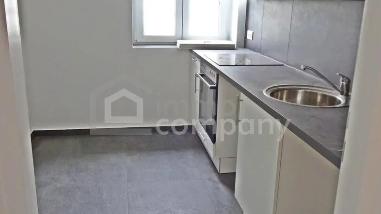 Moderne Küche mit weißen Schränken, grauer Arbeitsplatte, integriertem Backofen und Fliesenboden.