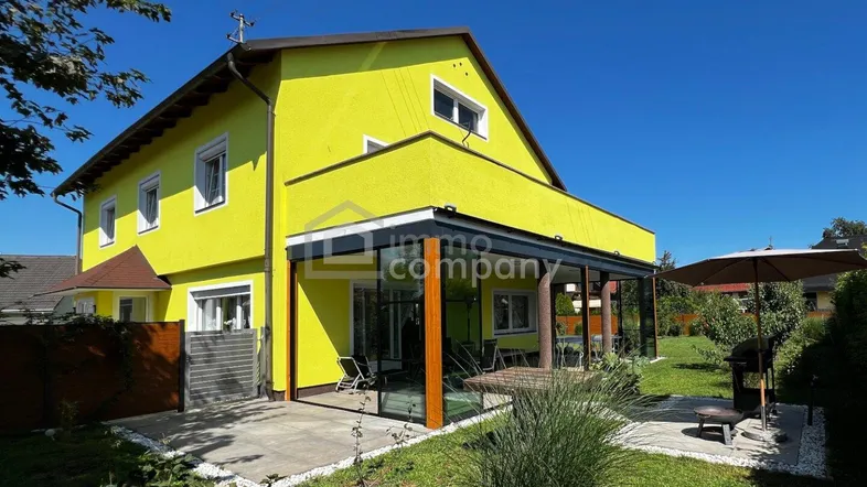 Gelbes Einfamilienhaus mit Wintergarten, Terrasse und gepflegtem Gartenbereich unter blauem Himmel.