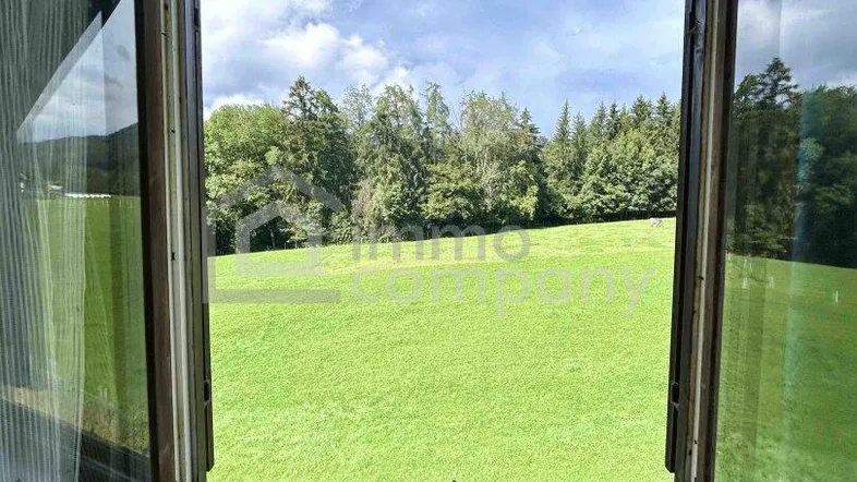 Blick aus einem geöffneten Fenster auf eine weite grüne Wiese und bewaldete Hügel unter bewölktem Himmel.