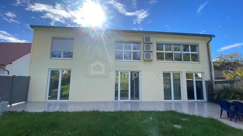 Hausrückseite mit großer Terrasse, Rasenfläche und Blick in den Garten unter blauem Himmel.