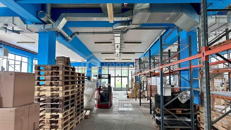 Geräumige Lagerhalle mit hohen Decken, blauen Stützbalken, Lüftungsrohren und Palettenregalen.