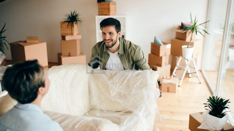 Zwei Personen tragen ein verpacktes Sofa in einem hellen Raum mit Umzugskartons, was auf einen Umzug hindeutet.