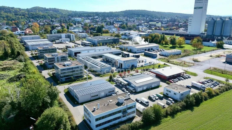 Luftaufnahme eines Gewerbegebiets mit verschiedenen Gebäuden, Parkplätzen und grünen Flächen unter blauem Himmel.
