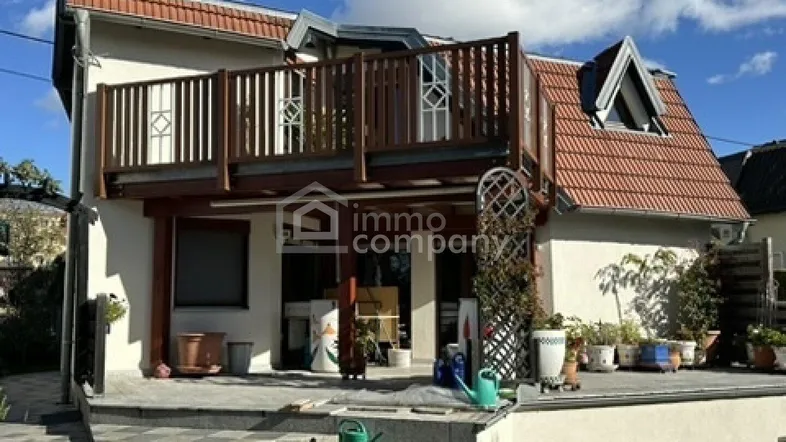 Hausfassade mit einer gepflasterten Terrasse im Erdgeschoss und einem darüberliegenden Holzbalkon.