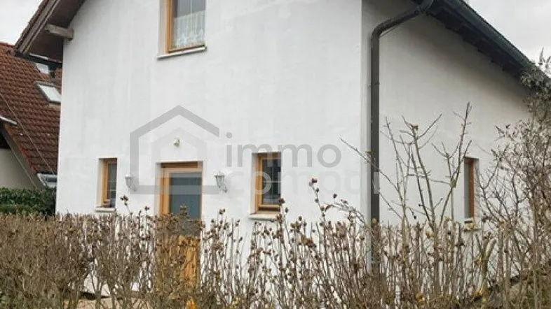 Seitenansicht des Hauses mit weißer Fassade, Holztür und gepflegter Hecke im Vordergrund.